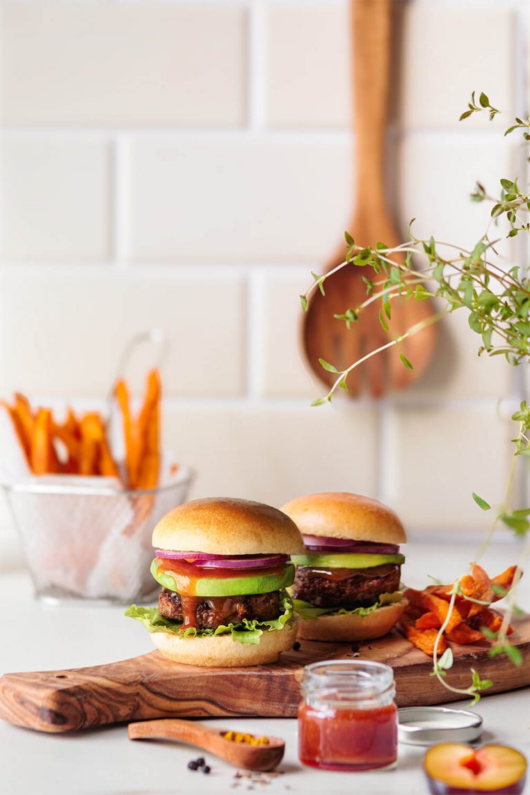 mini-burger mit avocado und scharfer pflaumensauce kleine burger mit avocado und pikanter pflaumensauce auf holzbrett, süßkartoffel-pommes im sieb, set in heller küche mit kacheln und holzlöffel – moderne, natürliche foodfotografie von natalia tag