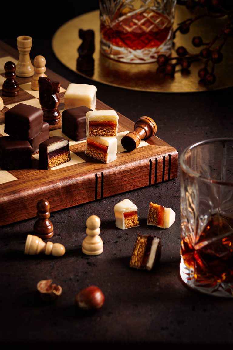 schachbrett mit dunklen und weißen dominosteinen, schachfiguren im fokus, whiskysgläser im anschnitt, dark & moody abendstimmung unter freunden – foodfotografie von natalia tag