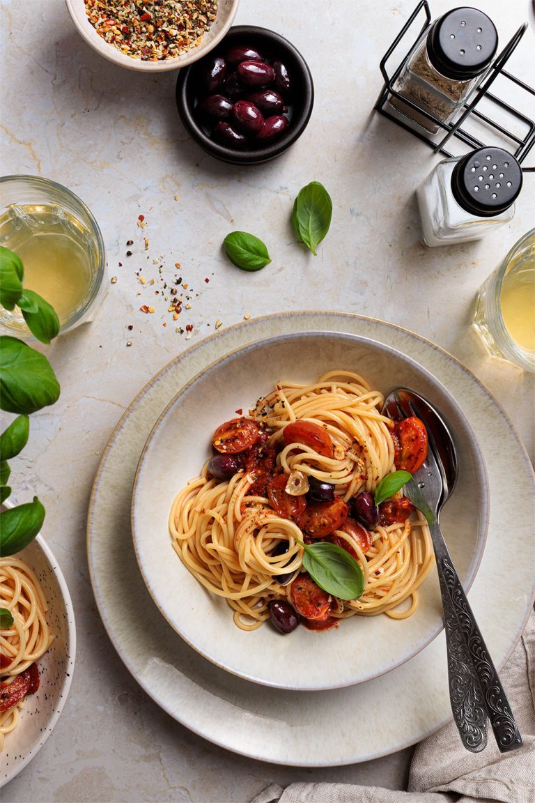 spaghetti mit Tomatensauce und kalamata-oliven, fotografiert im top-shot, modernes, klares styling, beige-weißer untergrund, gewürze und basilikumblätter auf dem Tisch – natürliche, klare foodfotografie von natalia tag